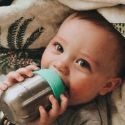 Baby holding a metallic cup with a green lid, lying on a patterned blanket.