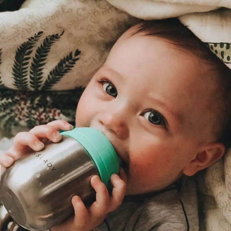 Baby holding a metallic cup with a green lid, lying on a patterned blanket.