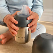 Child holding a stainless steel sippy cup with a silicone nipple on a wooden surface.